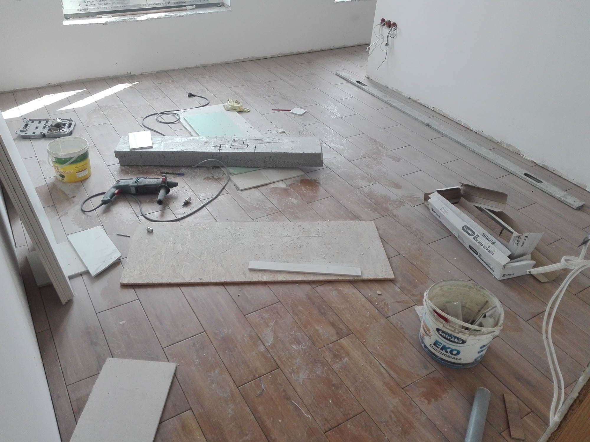 Podłoga z płytek imitujących drewno w trakcie układania, widoczne narzędzia, płyty OSB, poziomice oraz wiadro po farbie Śnieżka EKO.