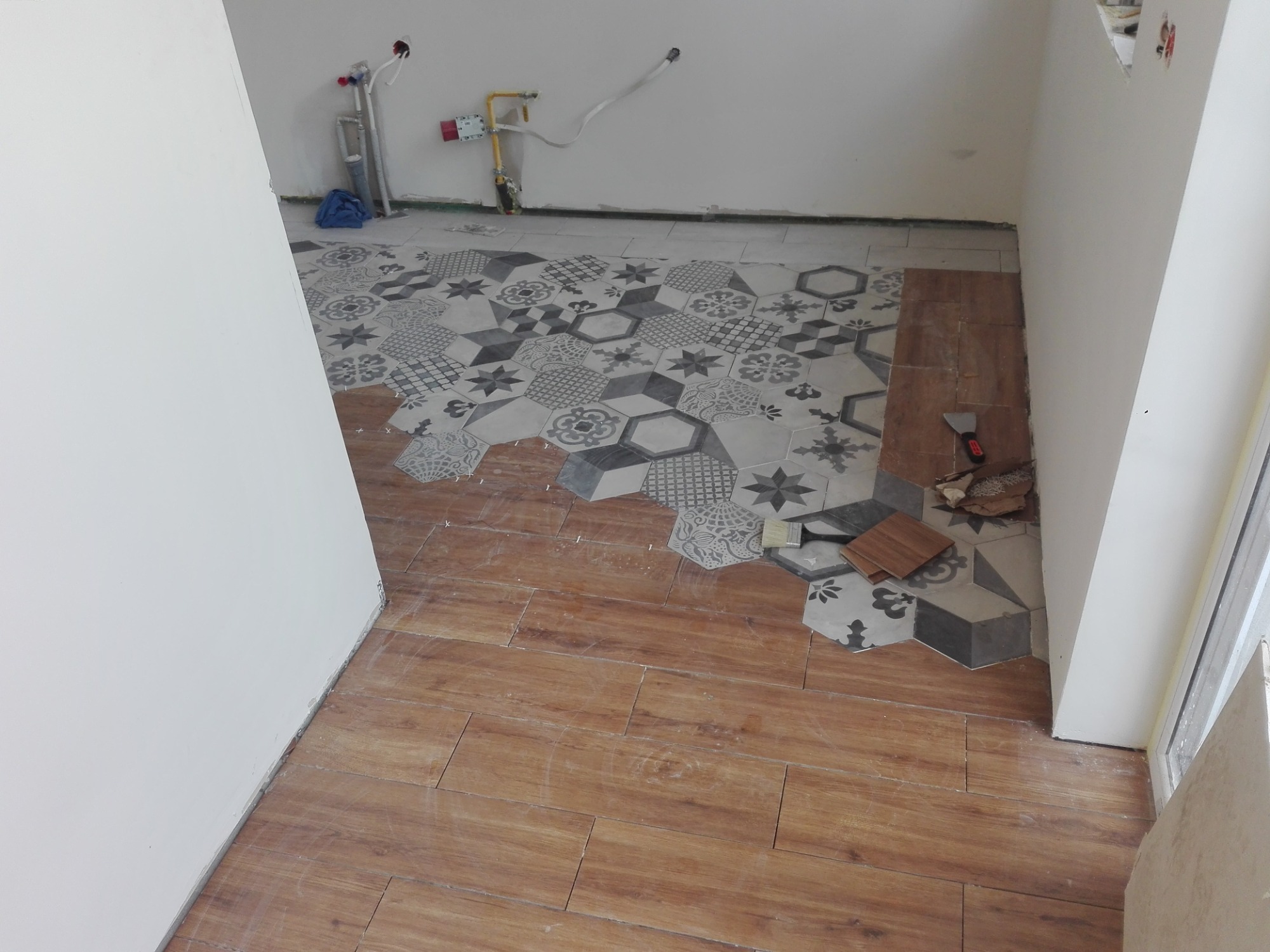Wnętrze w trakcie prac: połączenie heksagonalnych płytek podłogowych z wzorami i drewnopodobnych paneli, widoczne narzędzia i instalacje na ścianie.