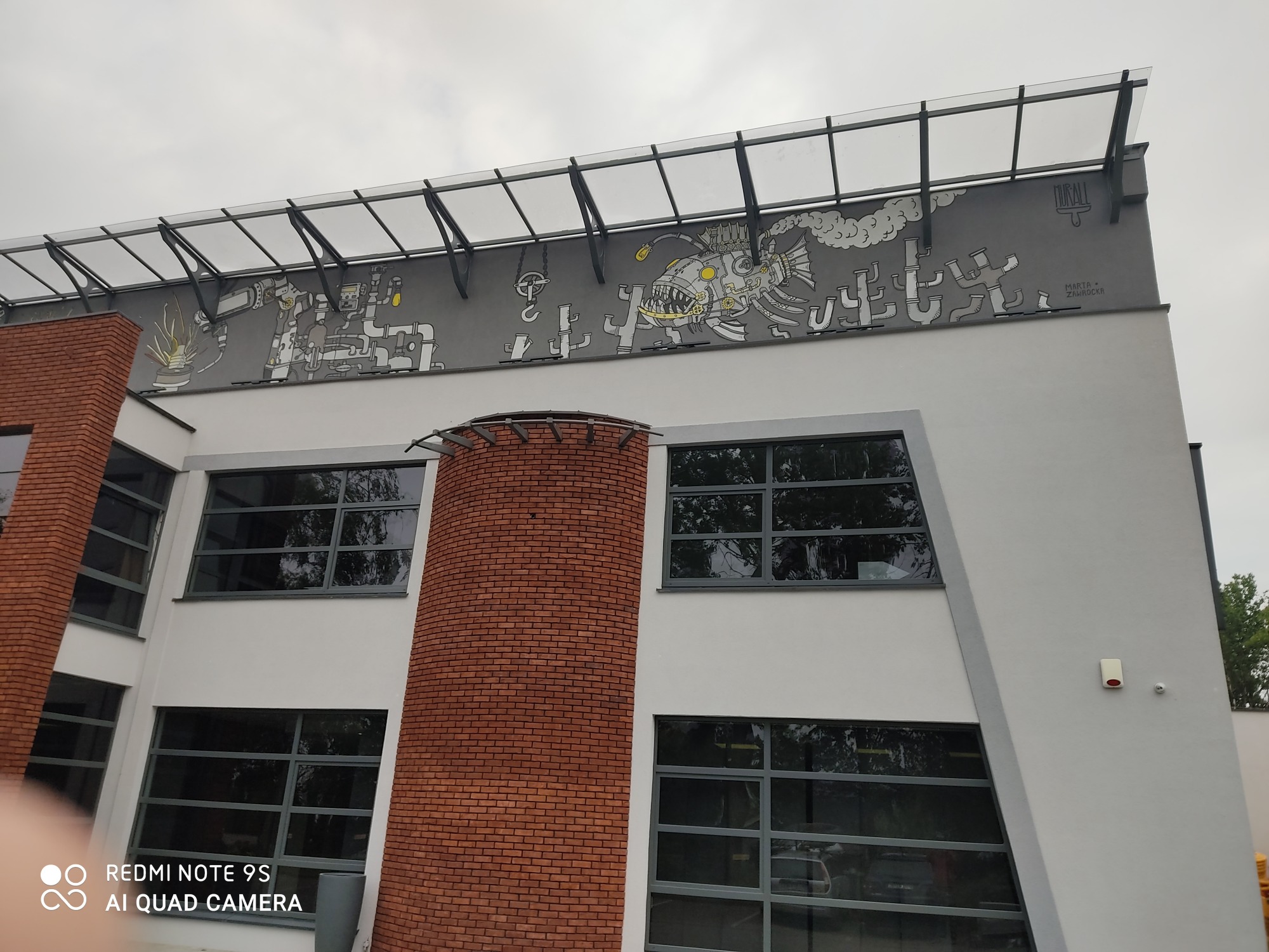 Zewnętrzny mural na budynku z motywami steampunkowymi, przedstawiający stylizowane maszyny i postacie, wykonany w odcieniach szarości i bieli z żółtymi akcentami, umieszczony pod zadaszeniem...