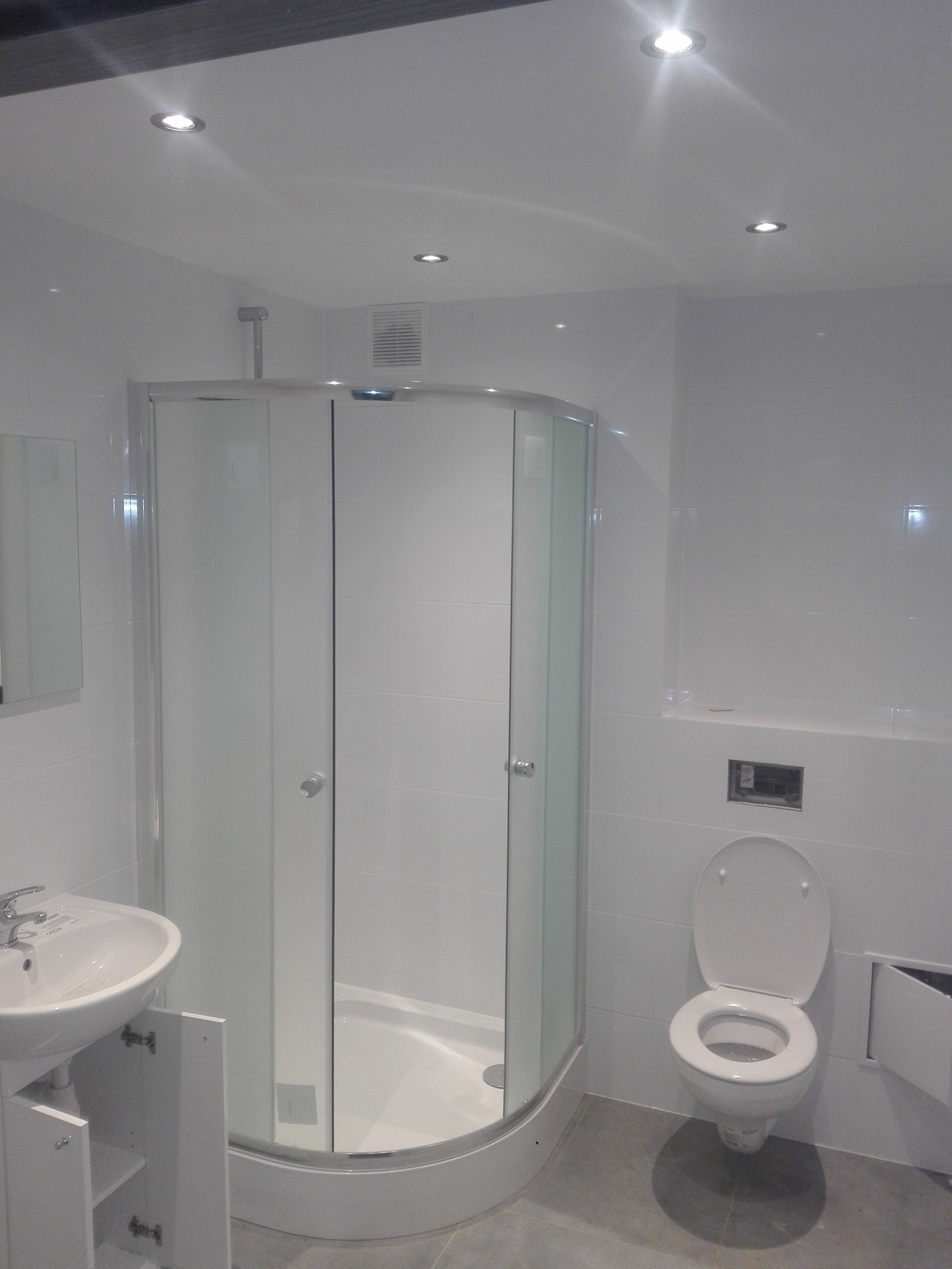 Nowoczesna łazienka z kabiną prysznicową narożną, białą umywalką z szafką i toaletą podwieszaną, oświetlona punktowymi lampami sufitowymi.