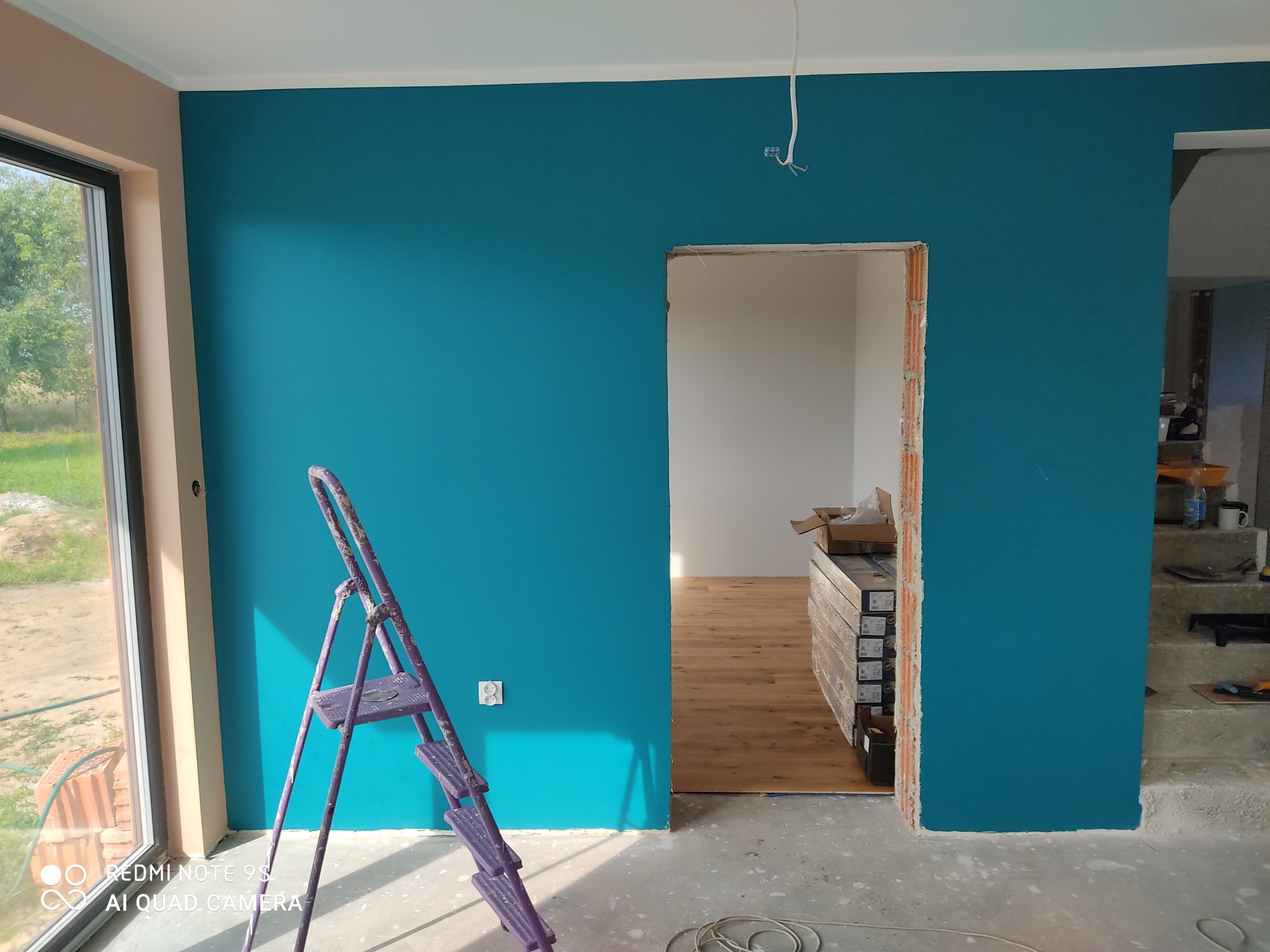 Pomieszczenie w trakcie malowania na intensywny turkusowy kolor, widoczna drabina malarska, świeżo pomalowana ściana, podłoga częściowo wyłożona panelami, surowy otwór drzwiowy i wystające kable...