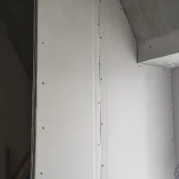 Ściana z płyt kartonowo-gipsowych zamontowana na metalowym stelażu, widoczne wkręty mocujące, surowy betonowy strop w tle.