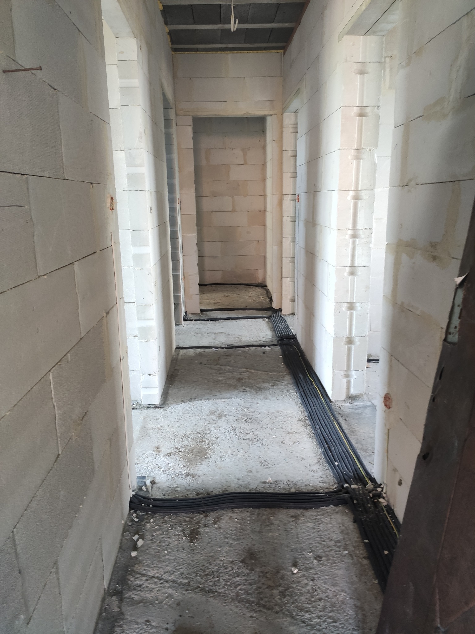 Korytarz w surowym stanie z widocznymi przewodami elektrycznymi ułożonymi na podłodze przed montażem podtynkowym, ściany z białych bloczków.