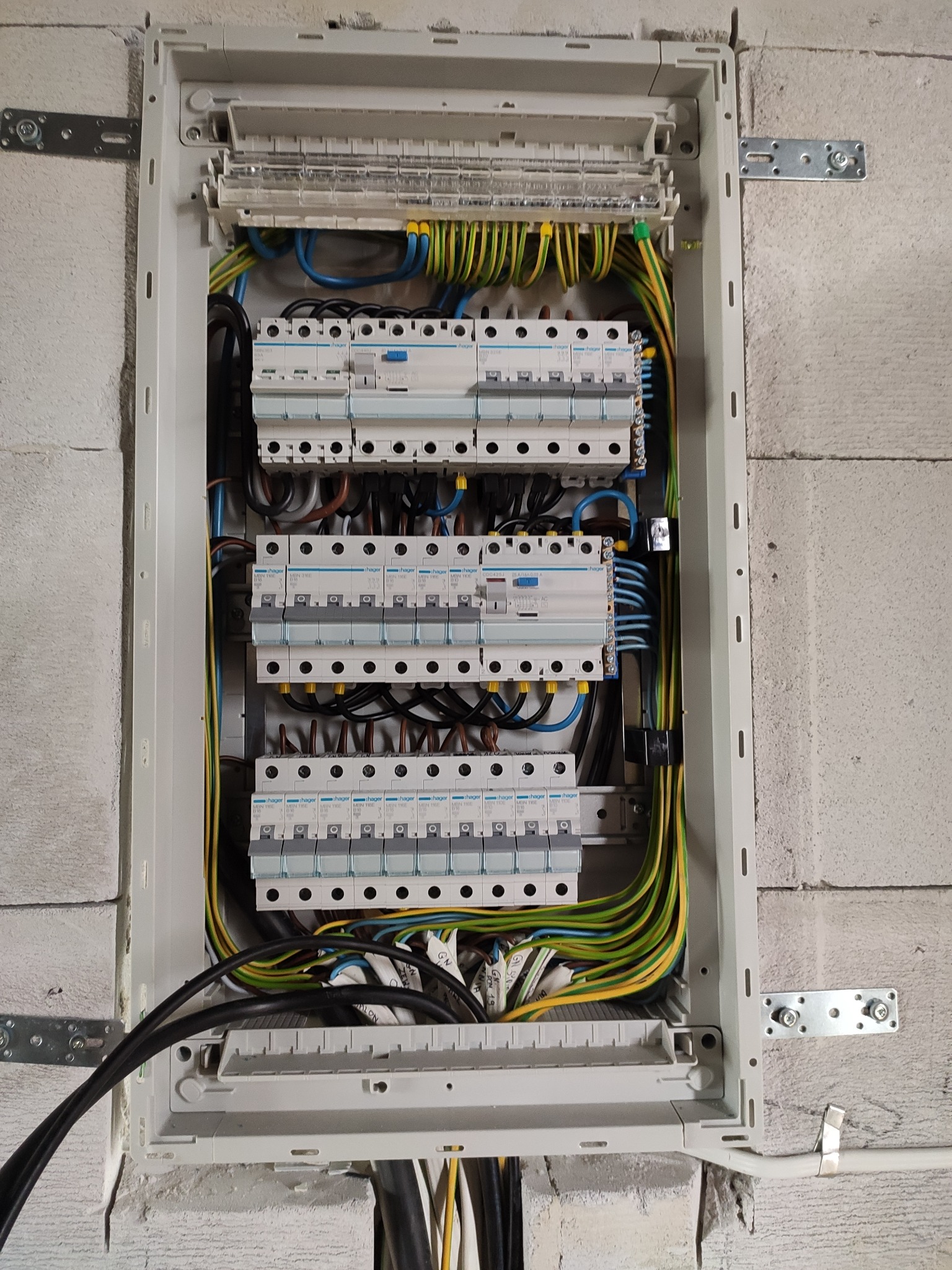 Rozdzielnia elektryczna z zamontowanymi bezpiecznikami Hager, widoczne przewody w kolorach żółto-zielonym, niebieskim i czarnym, zamontowana na ścianie z betonu komórkowego.