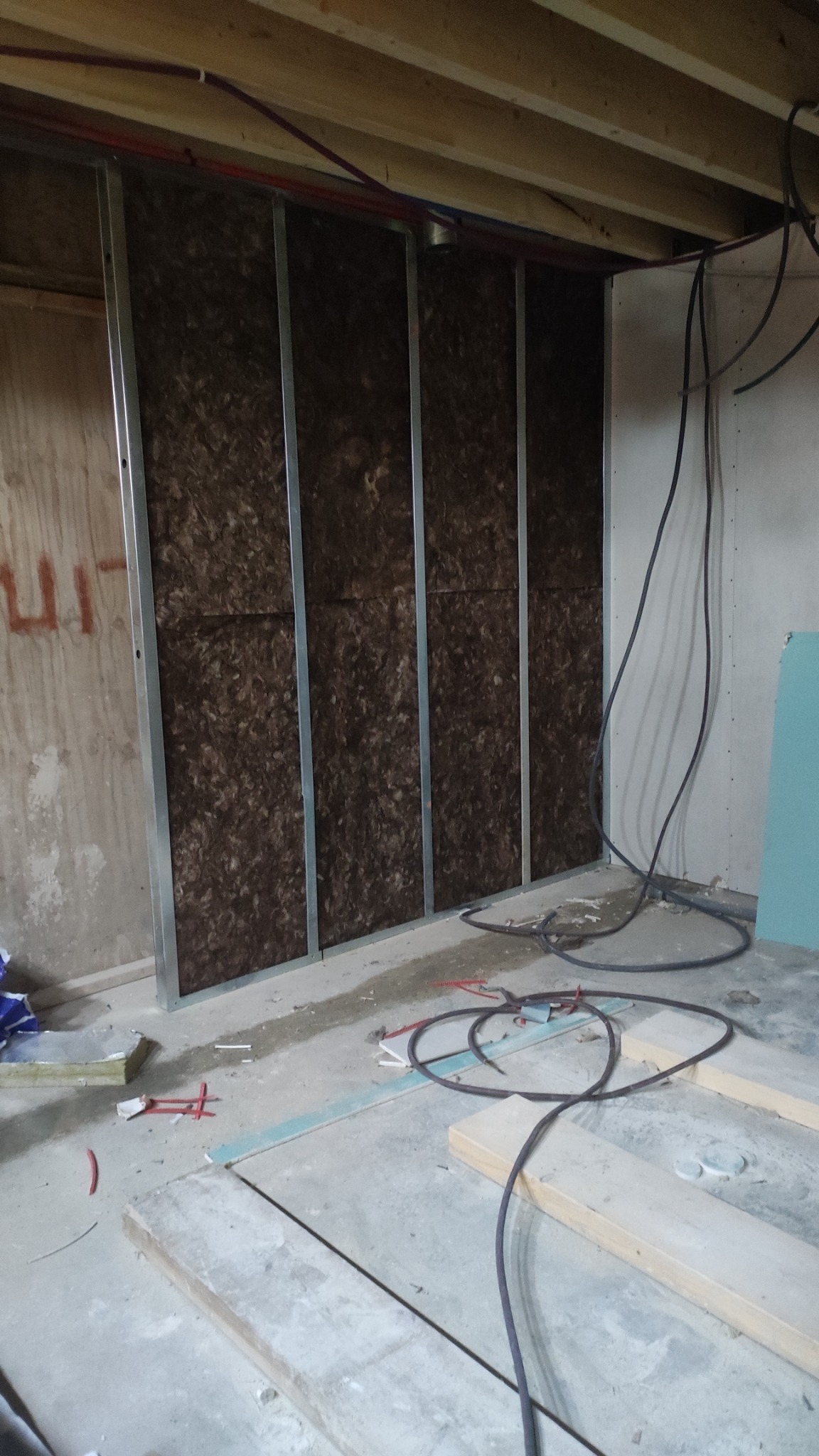 Montaż ściany z płyty karton gips z widoczną izolacją z wełny mineralnej pomiędzy metalowymi profilami, instalacje pod sufitem, surowy beton na podłodze, kable elektryczne w tle.