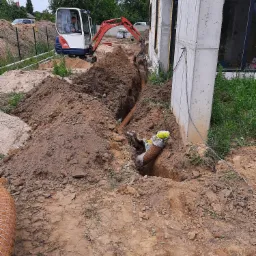 Mini koparka Kubota wykonuje wykop liniowy wzdłuż fundamentu budynku, widoczne rury kanalizacyjne w wykopie, oraz sterta ziemi.