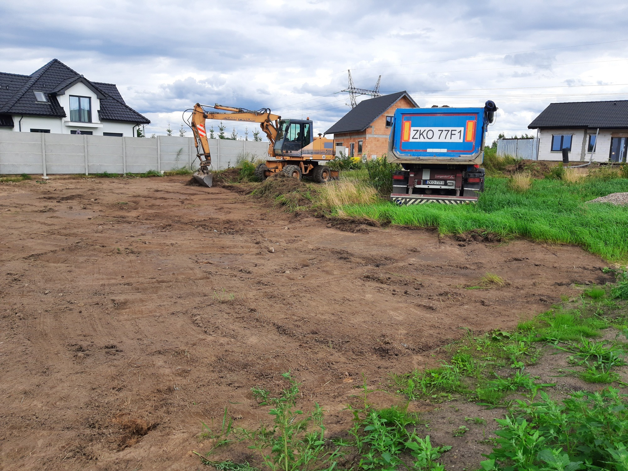 Żółta koparka i niebieska wywrotka na placu budowy, w tle domy jednorodzinne i pochmurne niebo.