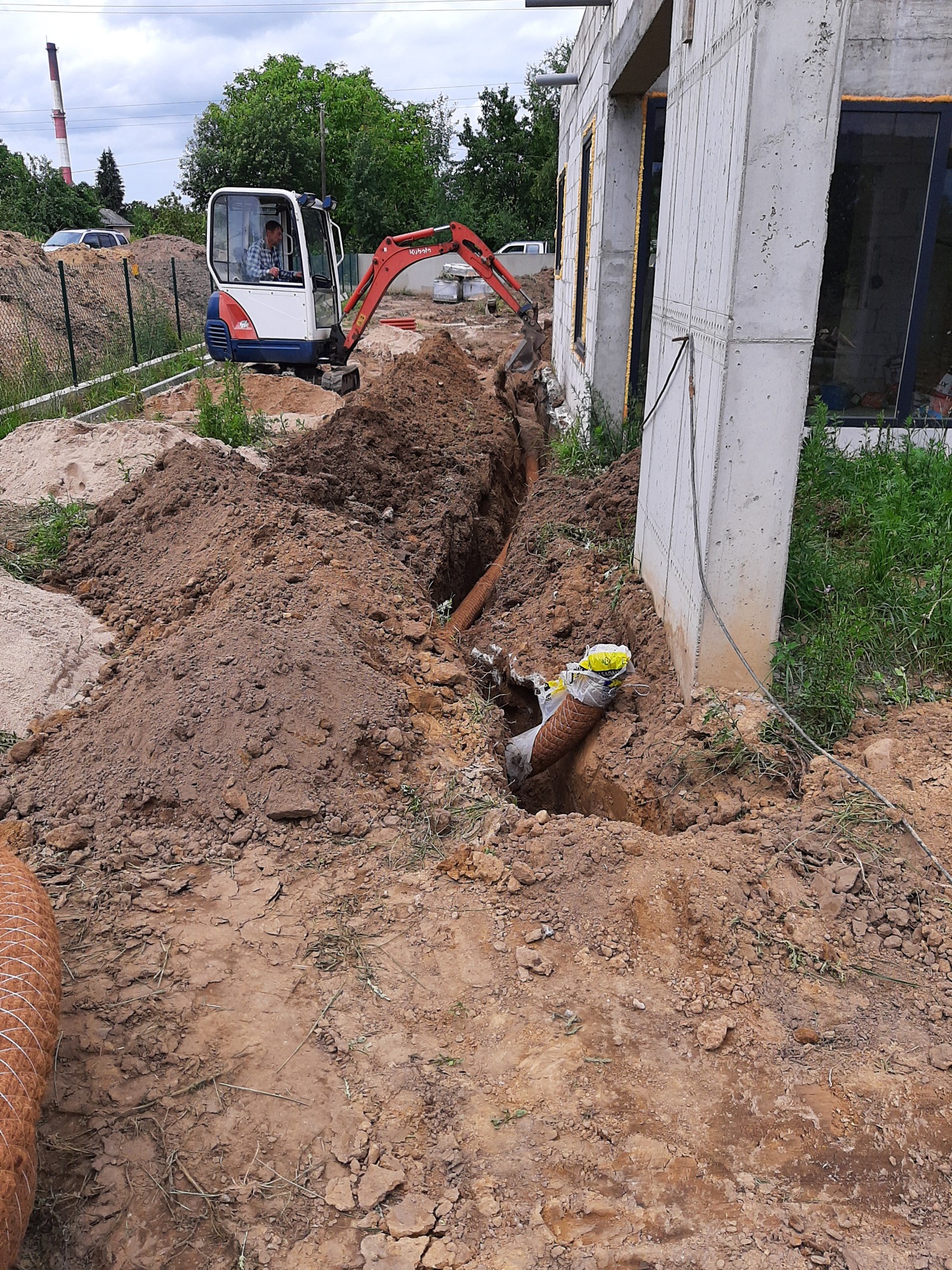 Mini koparka Kubota wykonuje wykop liniowy wzdłuż fundamentu budynku, widoczne rury kanalizacyjne w wykopie, oraz sterta ziemi.