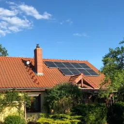 Czerwony dach z panelami słonecznymi i ceglanym kominem na tle błękitnego nieba z lekkimi chmurami, widok z poziomu ogrodu.