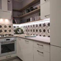 Biała kuchnia z błyszczącymi frontami, wzorzystymi płytkami na ścianie i drewnianymi półkami, oświetlona listwą LED pod szafkami wiszącymi, z wiankiem dekoracyjnym nad zlewem.