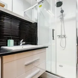 Nowoczesna łazienka z czarną, teksturowaną ścianą, białą umywalką nablatową z szafką z szufladami, kabiną prysznicową ze szklanymi drzwiami i wzorzystą podłogą.