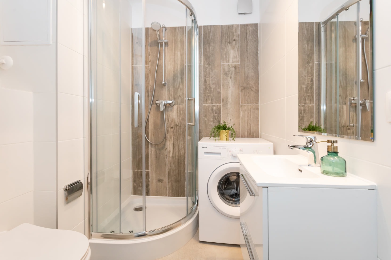 Jasna łazienka z kabiną prysznicową, umywalką z szafką i pralką Amica, drewnopodobne płytki na ścianie pod prysznicem, lustro odbijające kabinę