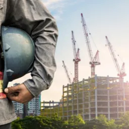 Pracownik w kamizelce odblaskowej trzyma kask na tle budowy z kilkoma żurawiami i budynkiem w tle.