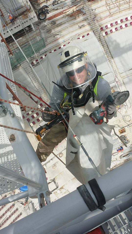 Pracownik w kasku i osłonie twarzy, zabezpieczony linami, używa szlifierki kątowej podczas pracy na wysokości na konstrukcji stalowej hali.