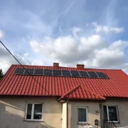 Panele słoneczne zamontowane na czerwonym dachu domu jednorodzinnego z numerem 11, widoczne kominy i błękitne niebo z chmurami w tle.