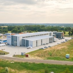 DXM Studio - Zdjęcie z lotu ptaka przedstawia nowoczesny budynek przemysłowy z szarego i białego metalu z logo firmy 'marbo' na fasadzie, otoczony parkingiem z samochodami i zielonym terenem.