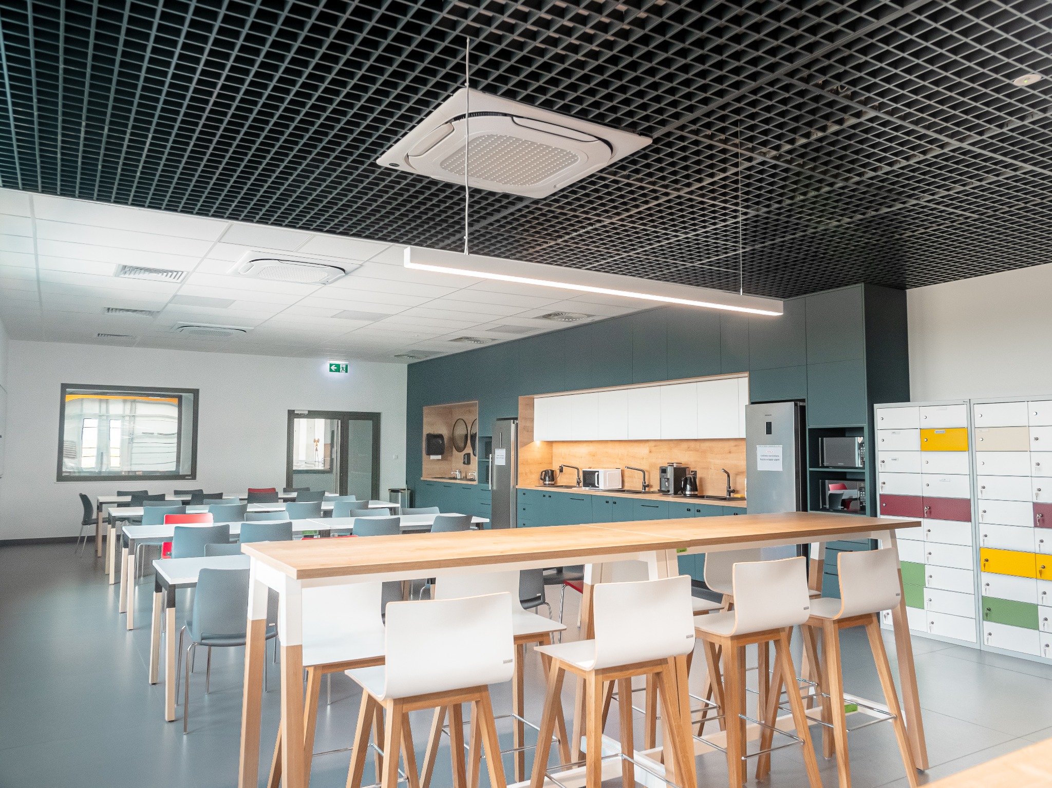 Nowoczesna przestrzeń biurowa z długim drewnianym stołem barowym, białymi hokerami, rzędami stolików z krzesłami i szafkami pracowniczymi w kolorach żółtym, zielonym i bordowym. Kuchnia z zabudową...