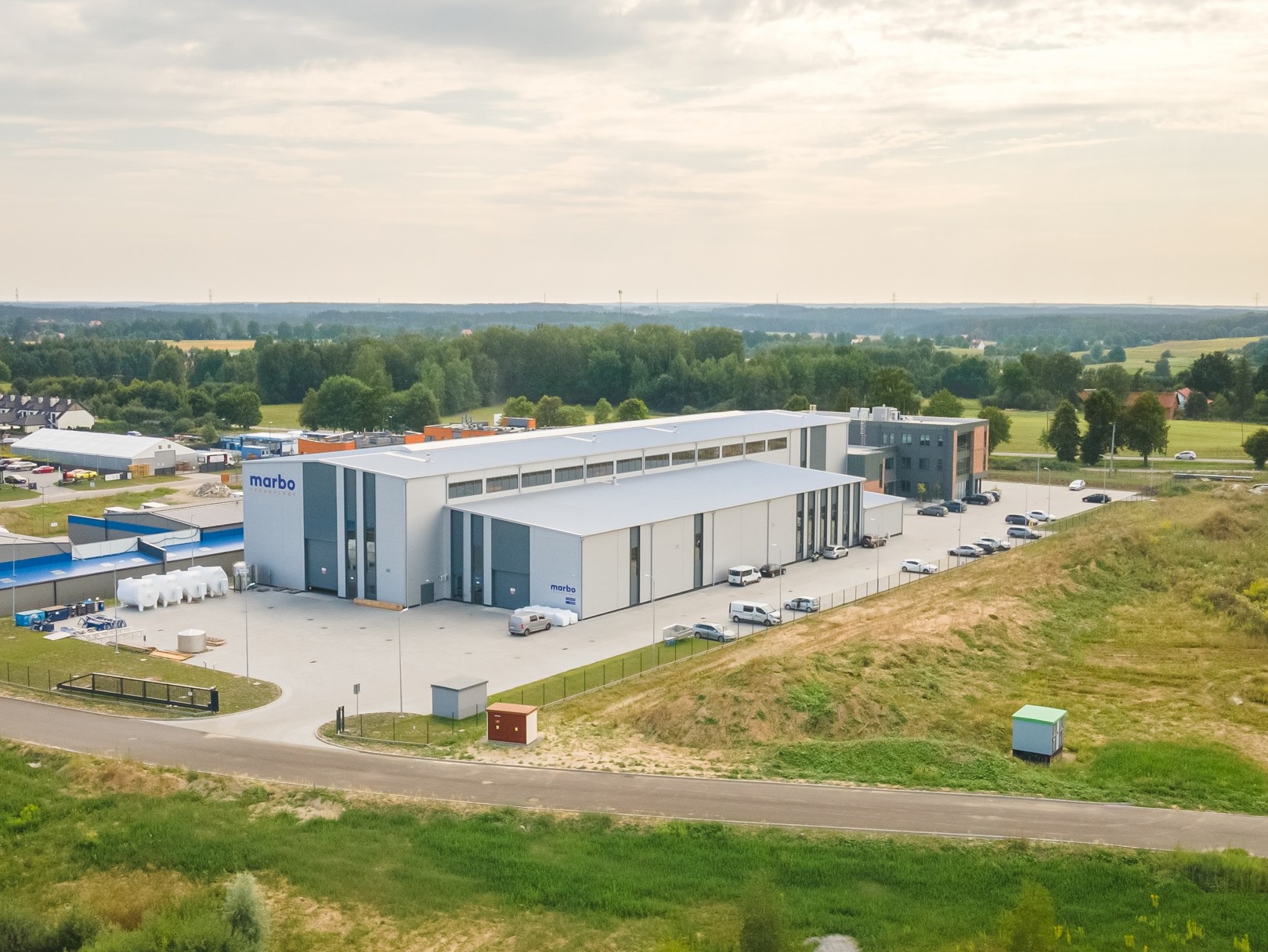 Zdjęcie z lotu ptaka przedstawia nowoczesny budynek przemysłowy z szarego i białego metalu z logo firmy 'marbo' na fasadzie, otoczony parkingiem z samochodami i zielonym terenem.