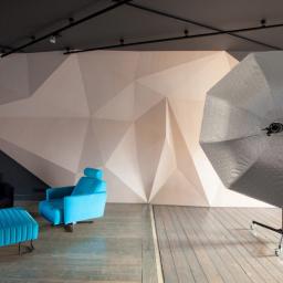 IN2IT Studio - Studio fotograficzne z geometryczną ścianą, niebieskim fotelem, czarnym fotelem i dużą srebrną parasolką oświetleniową.