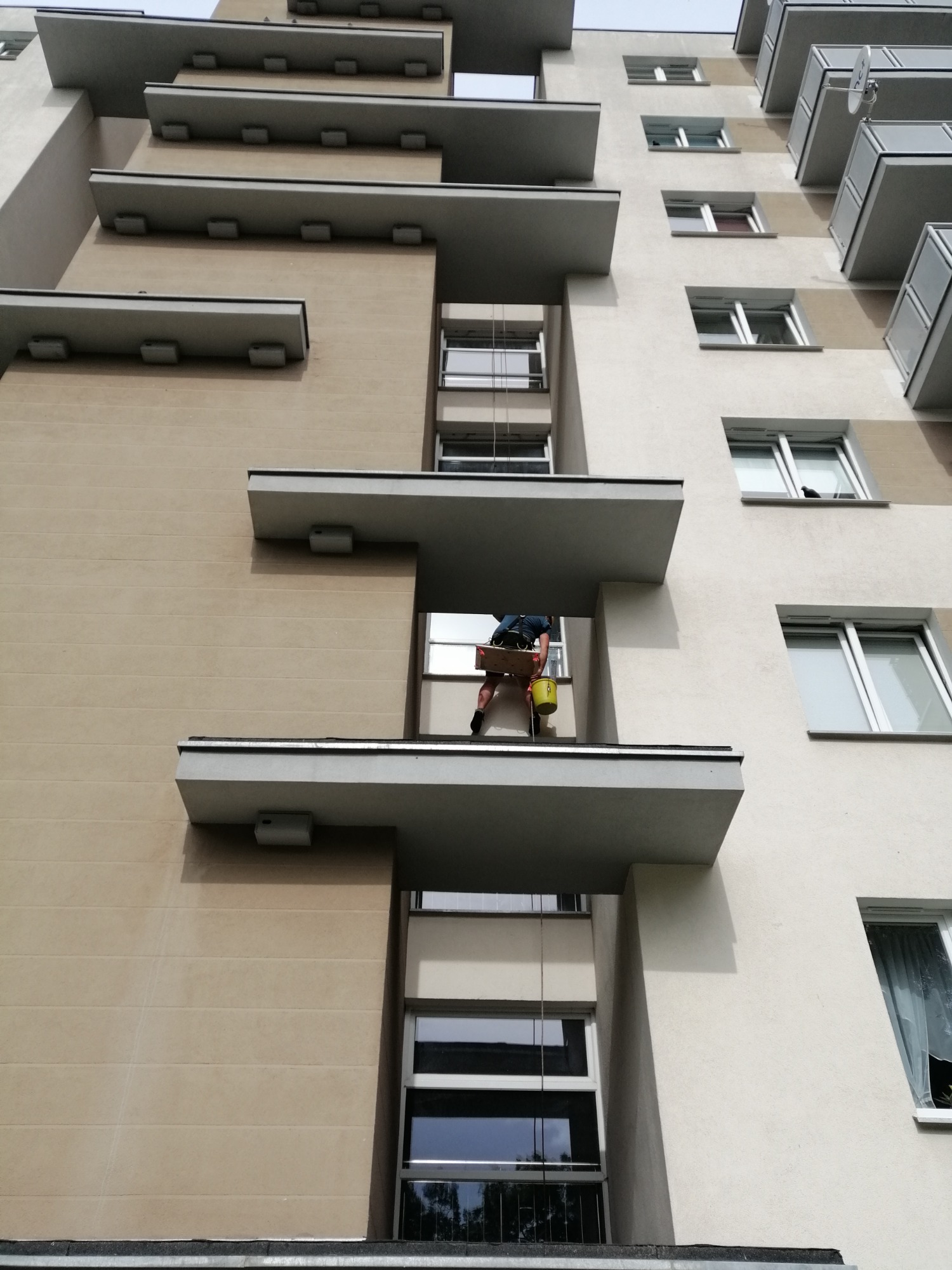 Pracownik na balkonie budynku wielorodzinnego, z żółtym wiadrem i skrzynką, prawdopodobnie podczas prac na wysokościach.