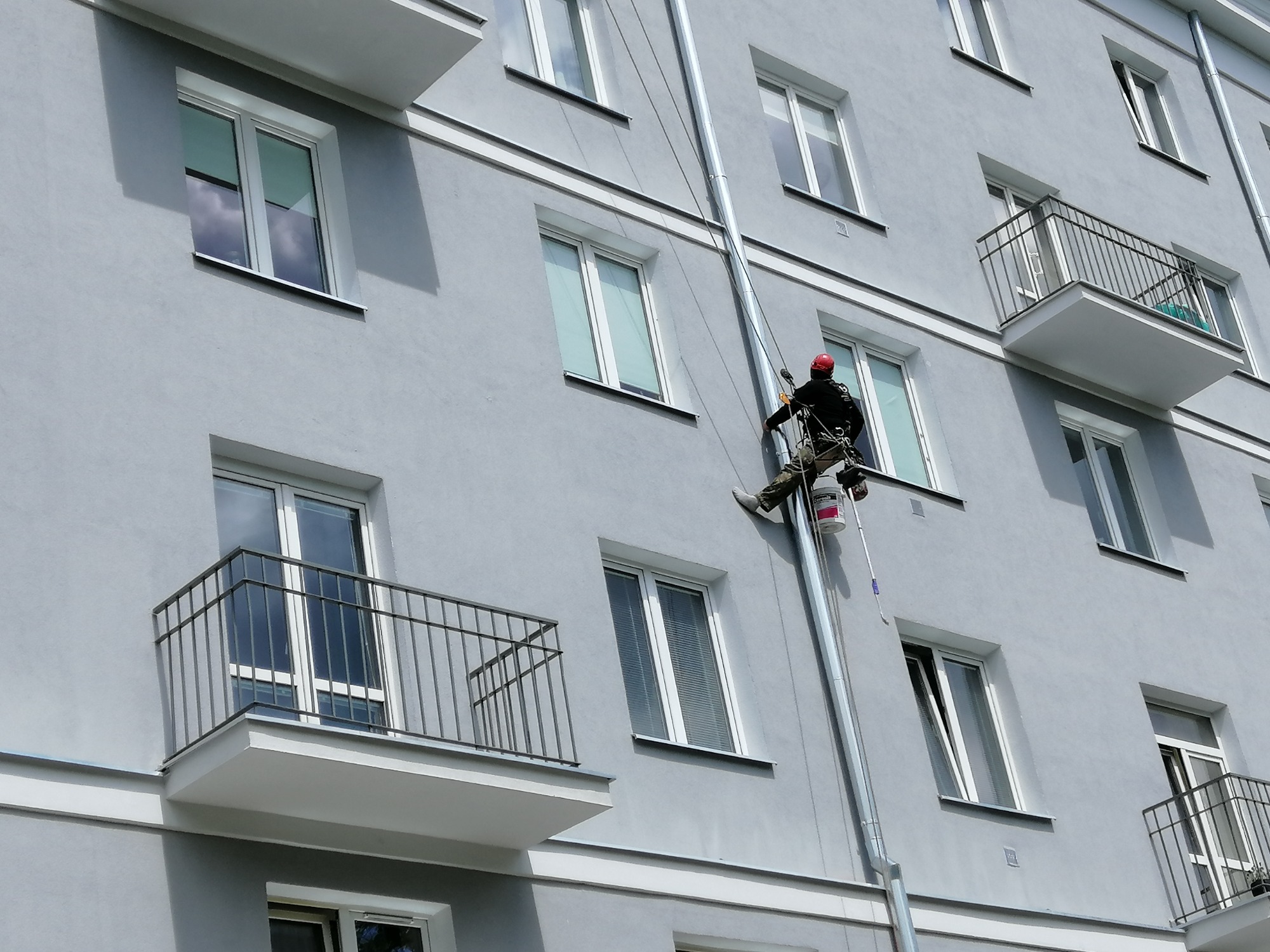 Pracownik na linach zabezpieczających, w czerwonym kasku, wykonuje prace na wysokości na szarej elewacji budynku z balkonami i oknami, widoczny kubełek z farbą marki Ceresit.