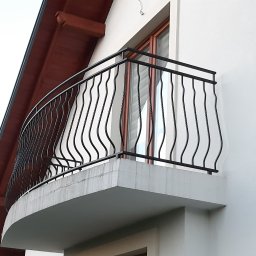 Usługi spawalnicze Custom-Metal Karol Lewicki - Czarna, metalowa balustrada balkonowa z falistymi prętami, zamontowana na białym, półokrągłym balkonie przy białej ścianie budynku z drewnianym okapem.