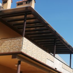Usługi spawalnicze Custom-Metal Karol Lewicki - Zadaszenie balkonu z brązową metalową konstrukcją i przezroczystym dachem, widoczne z dołu na tle błękitnego nieba.