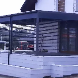 Nowoczesna, szara konstrukcja z metalowymi lamelami, prawdopodobnie zadaszenie tarasu, przylegająca do białego budynku z drewnianymi żaluzjami.