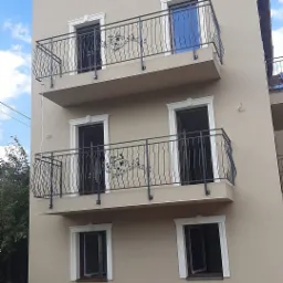 Elewacja budynku z beżową farbą, dwa balkony z czarnymi, metalowymi balustradami z motywem roślinnym, okrągłe okno na poddaszu i stosy kostki brukowej na ziemi.