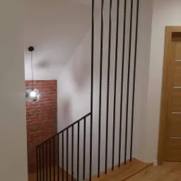 Drewniane schody z czarną, metalową balustradą i pionowymi prętami zabezpieczającymi, uzupełnione ceglaną ścianą i nowoczesną lampą wiszącą.