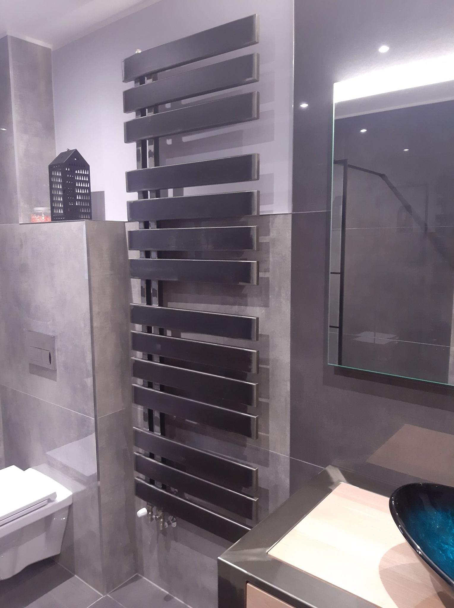 Szary grzejnik łazienkowy z poziomych, prostokątnych profili zamontowany na ścianie w łazience w Przemyślu, widok z boku na umywalkę i toaletę.