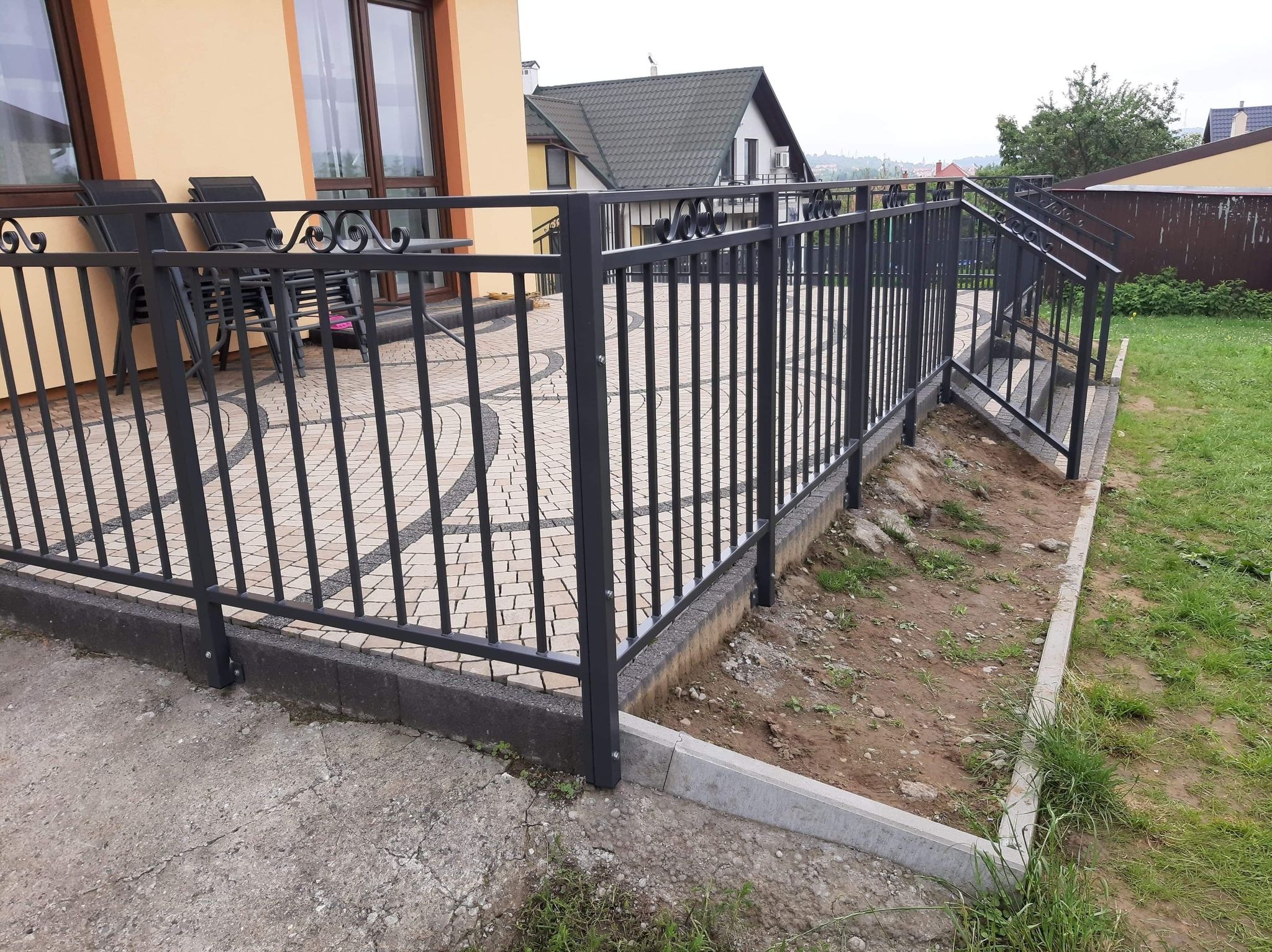 Zewnętrzna, czarna barierka z pionowych prętów i ozdobnych elementów na górze, oddzielająca taras z kostki brukowej od trawnika i schodów.