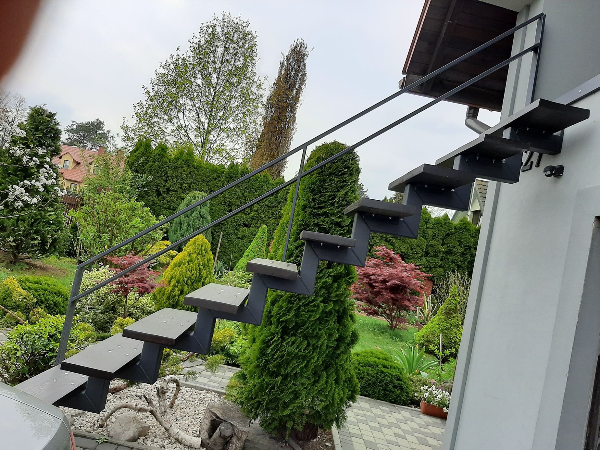 Zewnętrzne schody z metalową konstrukcją i drewnianymi stopniami, prowadzące do domu, otoczone bujną zielenią ogrodu.
