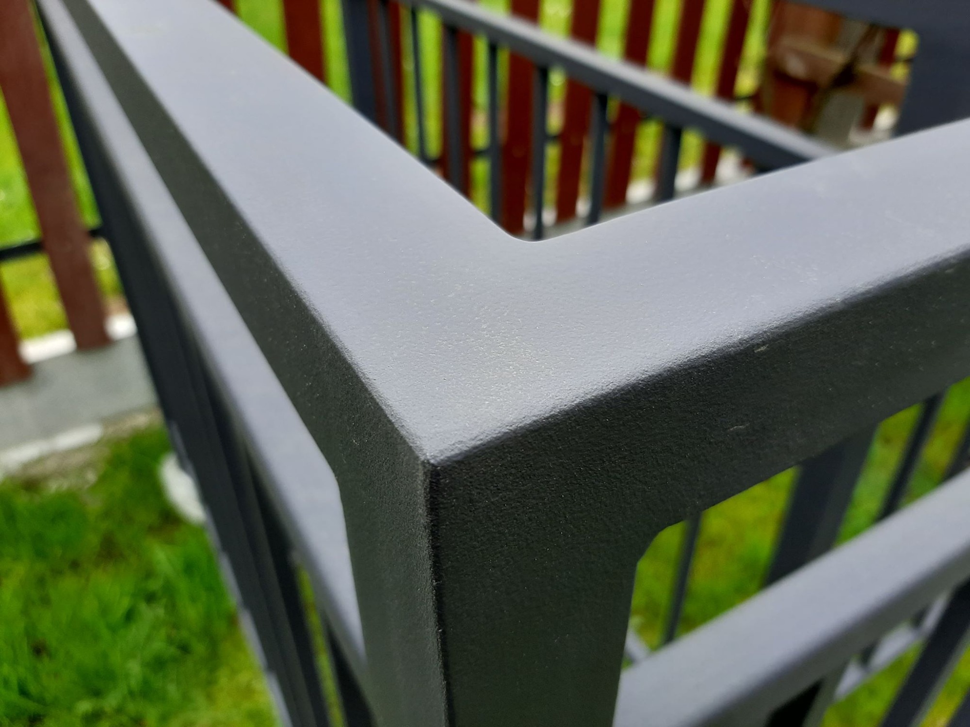 Fragment metalowej konstrukcji balustrady pomalowanej na grafitowo, widoczny narożnik i pionowe elementy, tło stanowi trawa i elementy ogrodzenia.