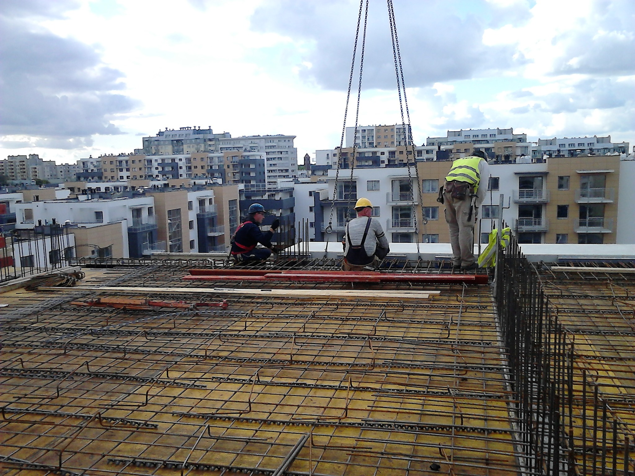 Trzech robotników w kaskach i kamizelce odblaskowej pracuje na zbrojeniu stropu budynku wielorodzinnego, w tle widoczne bloki mieszkalne i pochmurne niebo.