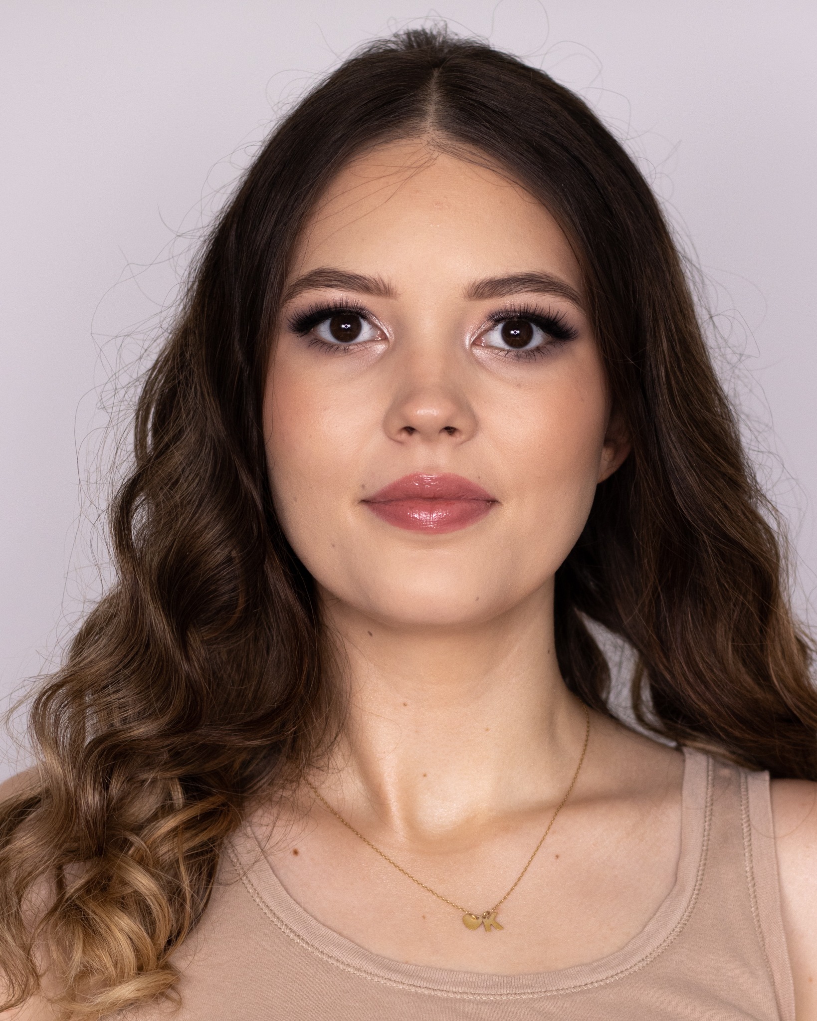 Portret młodej kobiety z profesjonalnym makijażem, podkreślone oczy z długimi rzęsami i błyszczące usta, delikatna biżuteria na szyi.