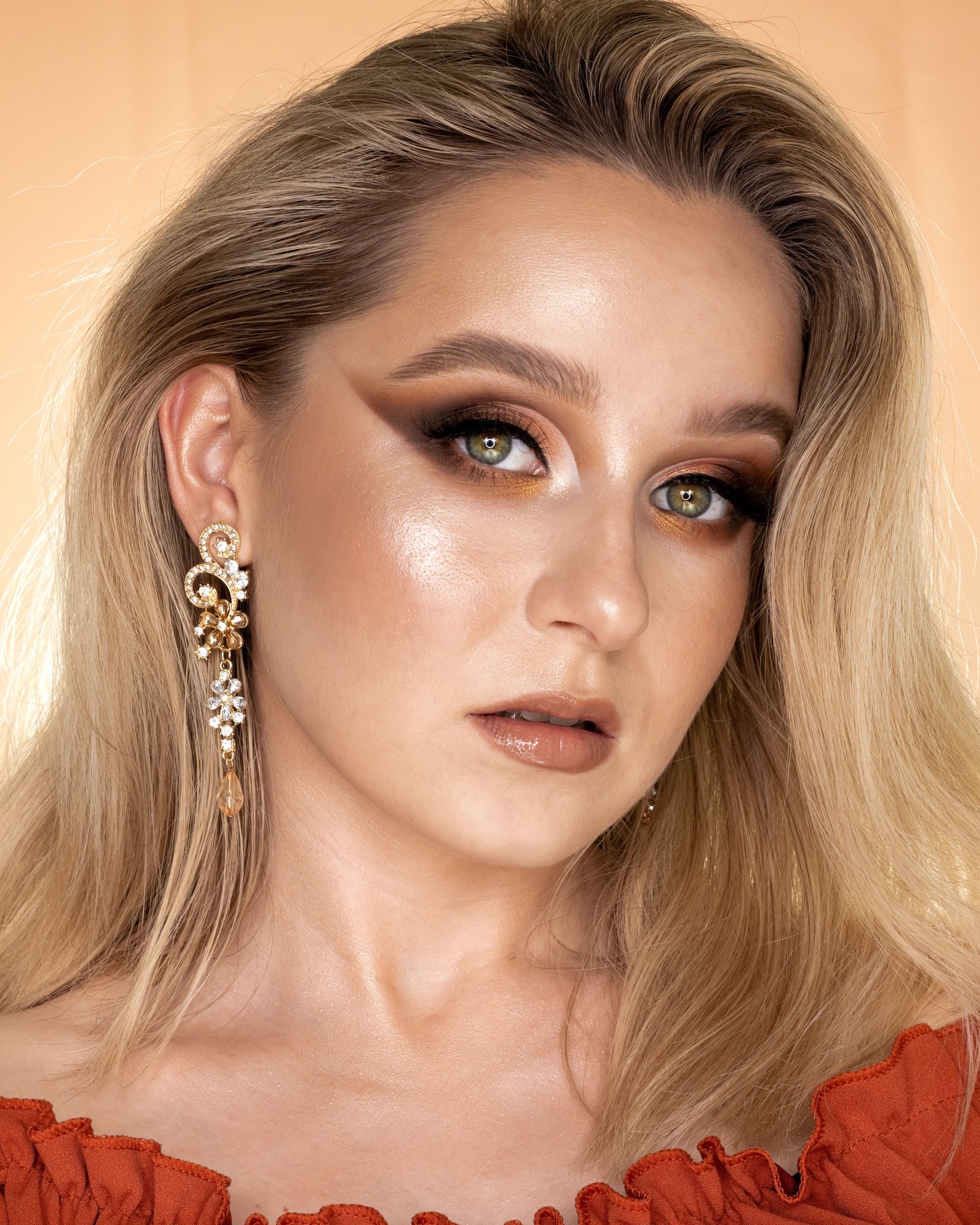 Portret kobiety z blond włosami i wyrazistym, brązowo-złotym cieniowaniem powiek, błyszczącą cerą i błyszczykiem na ustach, z długim, bogato zdobionym kolczykiem z kryształkami.