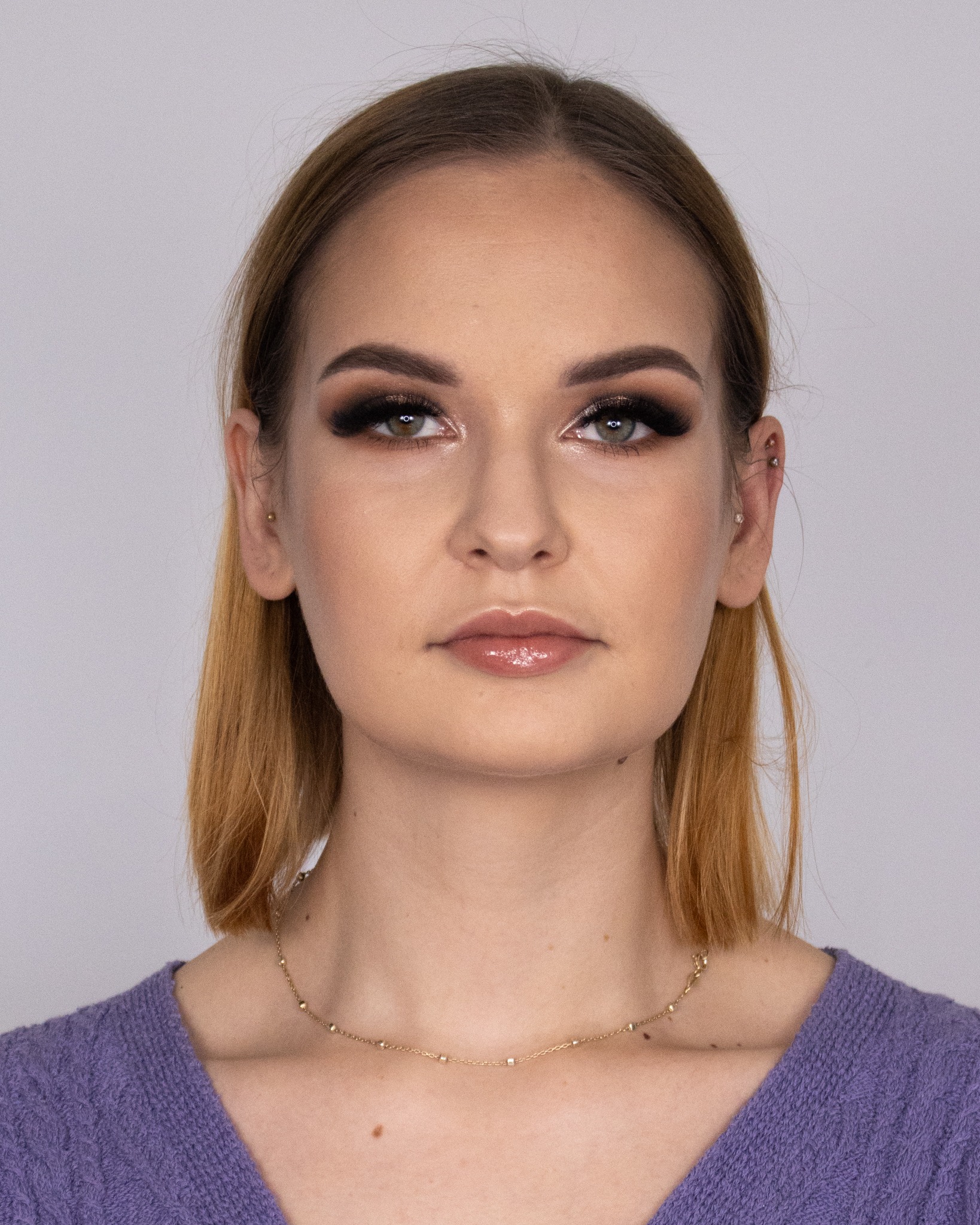 Portret kobiety z profesjonalnym makijażem oczu w odcieniach brązu i czerni, błyszczącymi ustami i złotym naszyjnikiem na tle szarego tła.