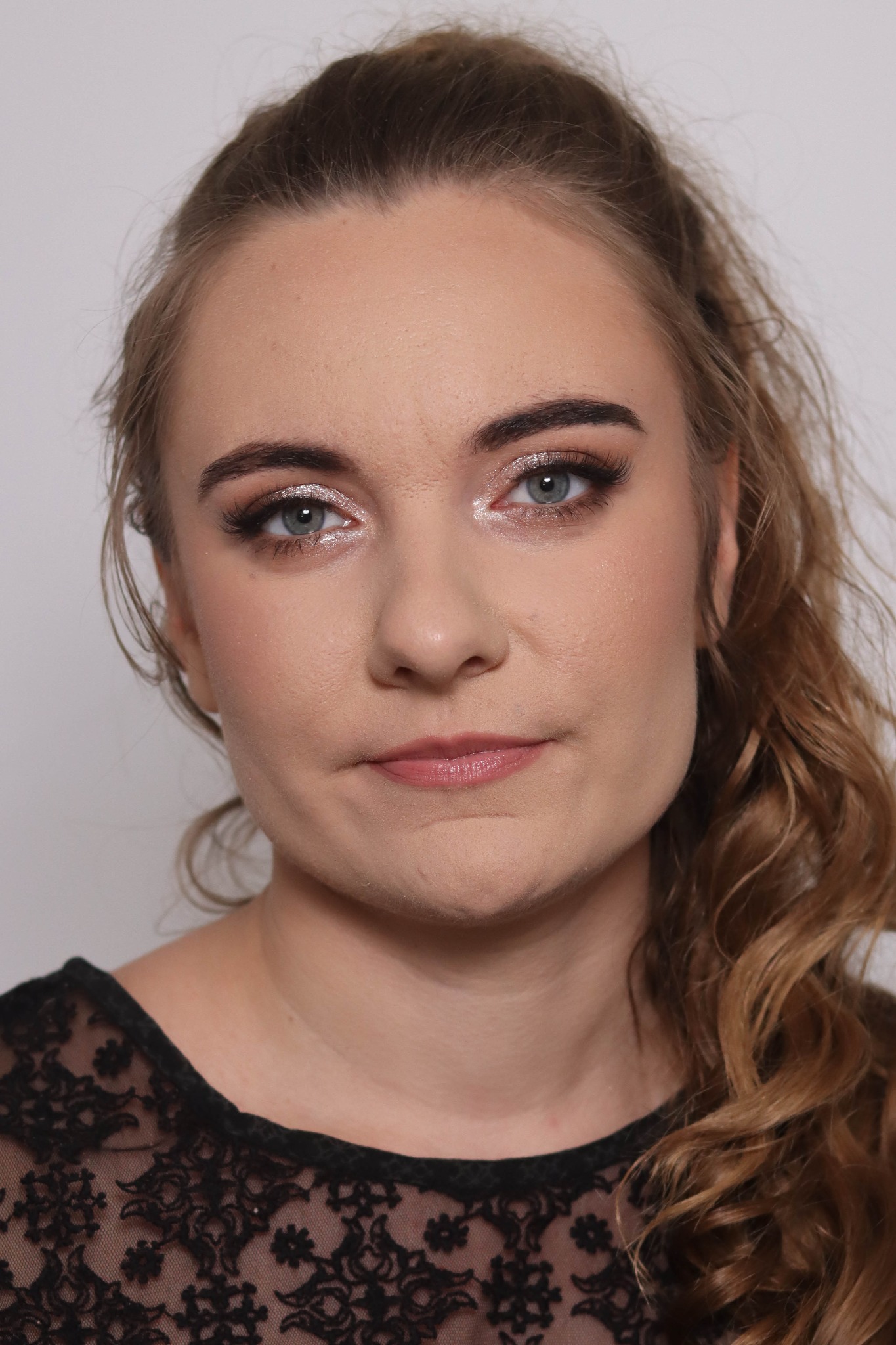 Portret kobiety z delikatnym, błyszczącym cieniem na powiekach i podkreślonymi rzęsami, włosy upięte z boku w luźne fale, ubrana w czarną koronkową bluzkę.