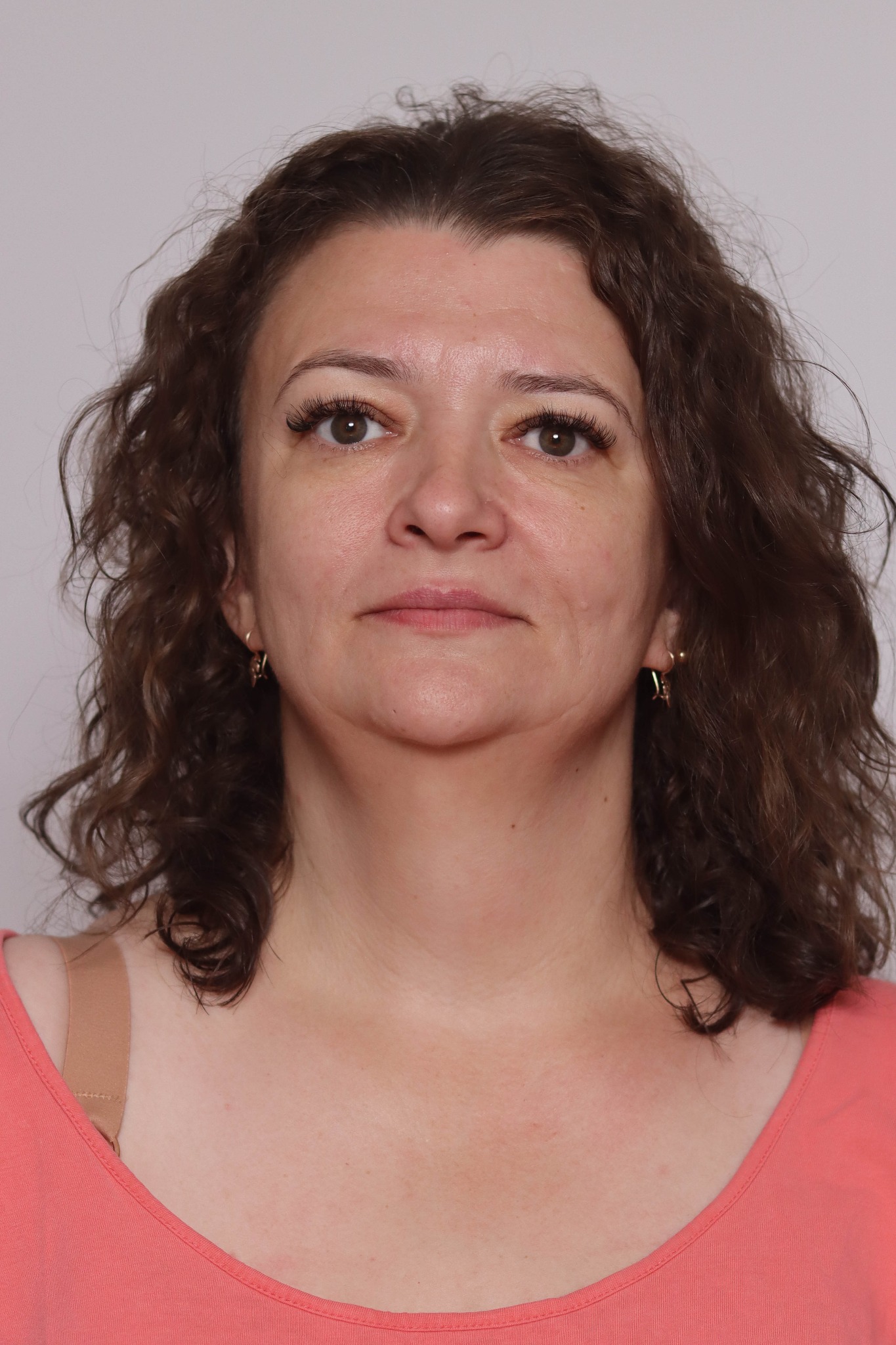 Portret kobiety z brązowymi kręconymi włosami i przedłużonymi rzęsami, ubrana w koralową koszulkę, na jasnym tle.