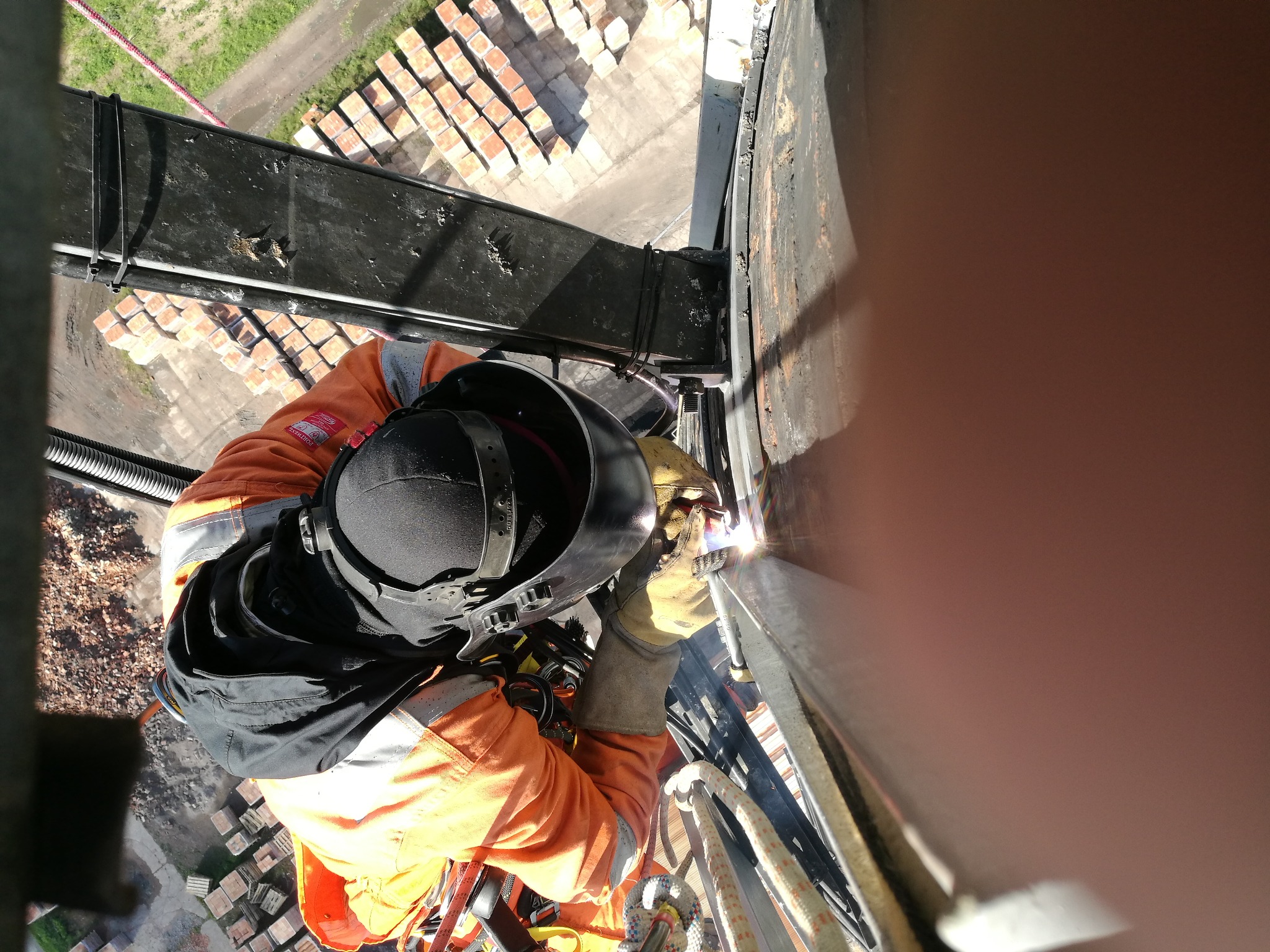 Spawacz w pomarańczowym kombinezonie na wysokości, spawający metalową konstrukcję z widokiem na cegły i teren.