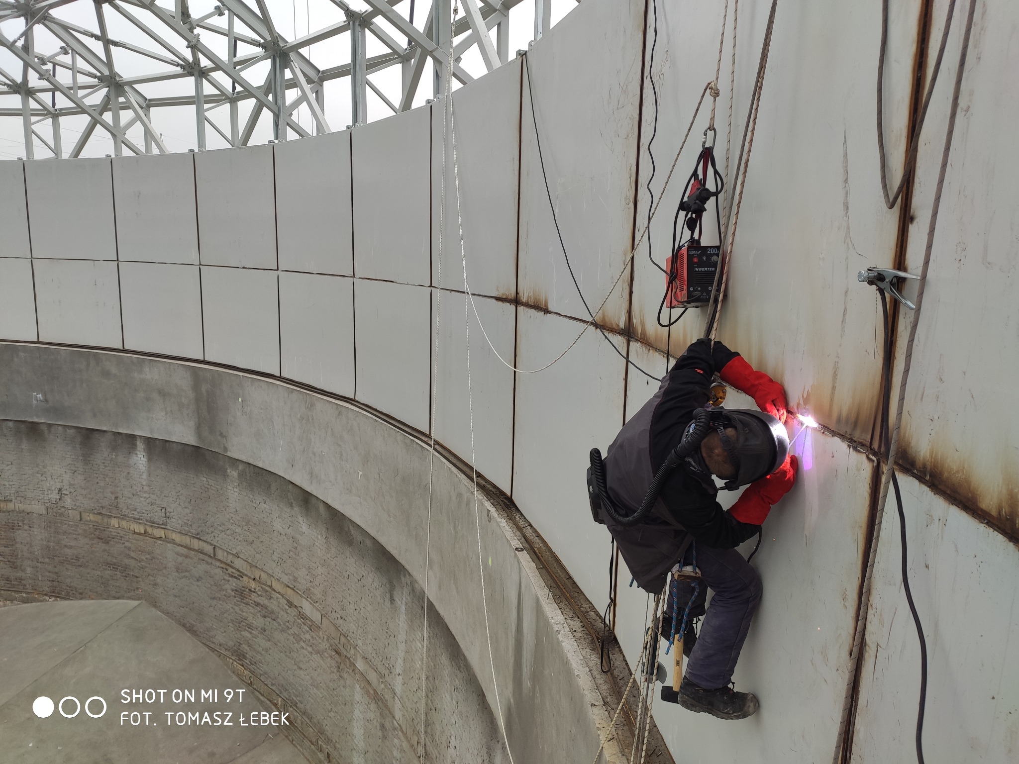 Spawacz w kasku i czerwonych rękawicach, zabezpieczony linami, spawa metalową konstrukcję na wysokości, widoczna spawarka i iskry.
