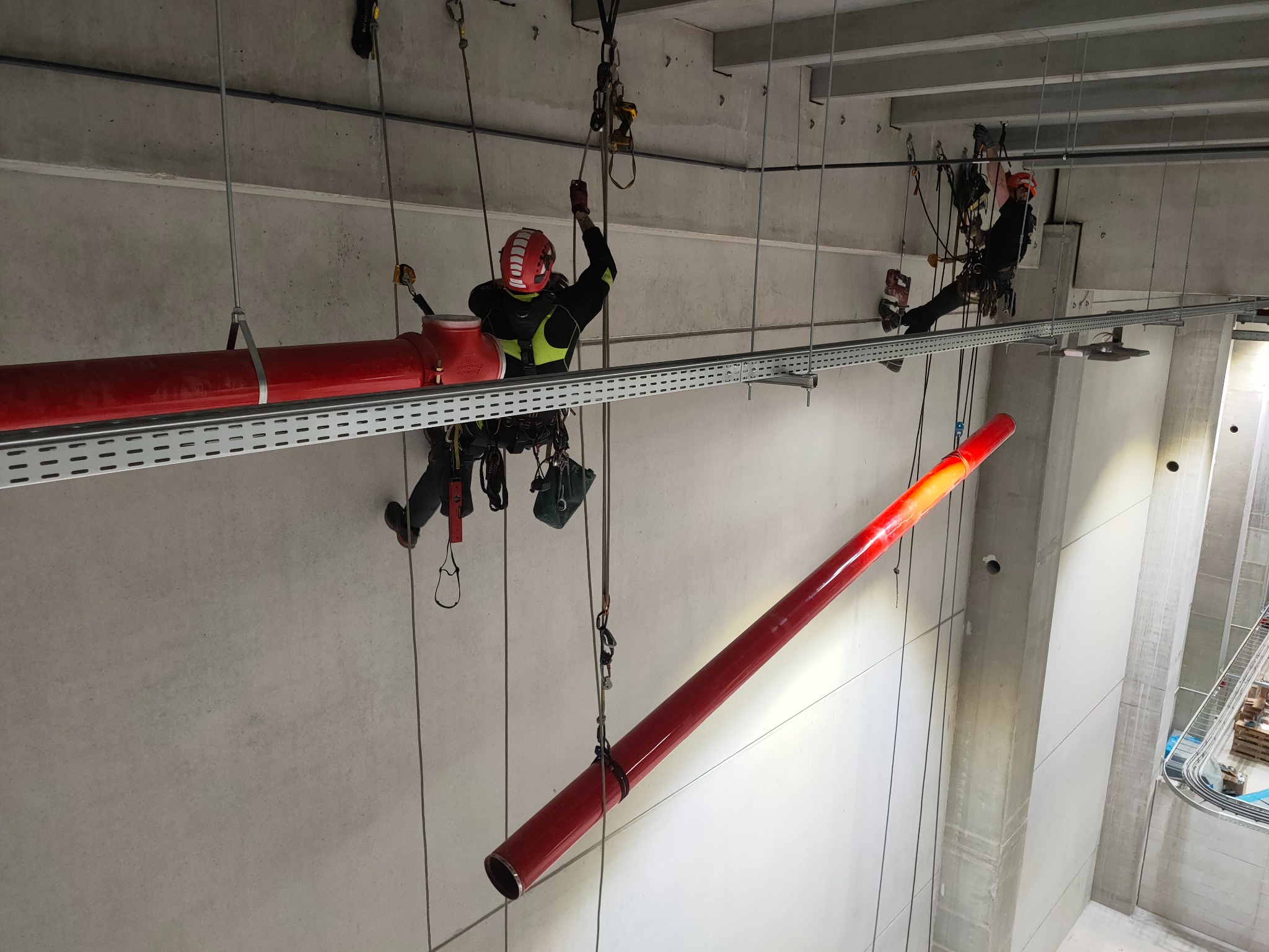 Dwóch pracowników na wysokościach, zabezpieczonych linami, montuje czerwoną rurę pod sufitem hali przemysłowej. Widoczne stalowe konstrukcje podtrzymujące instalację.