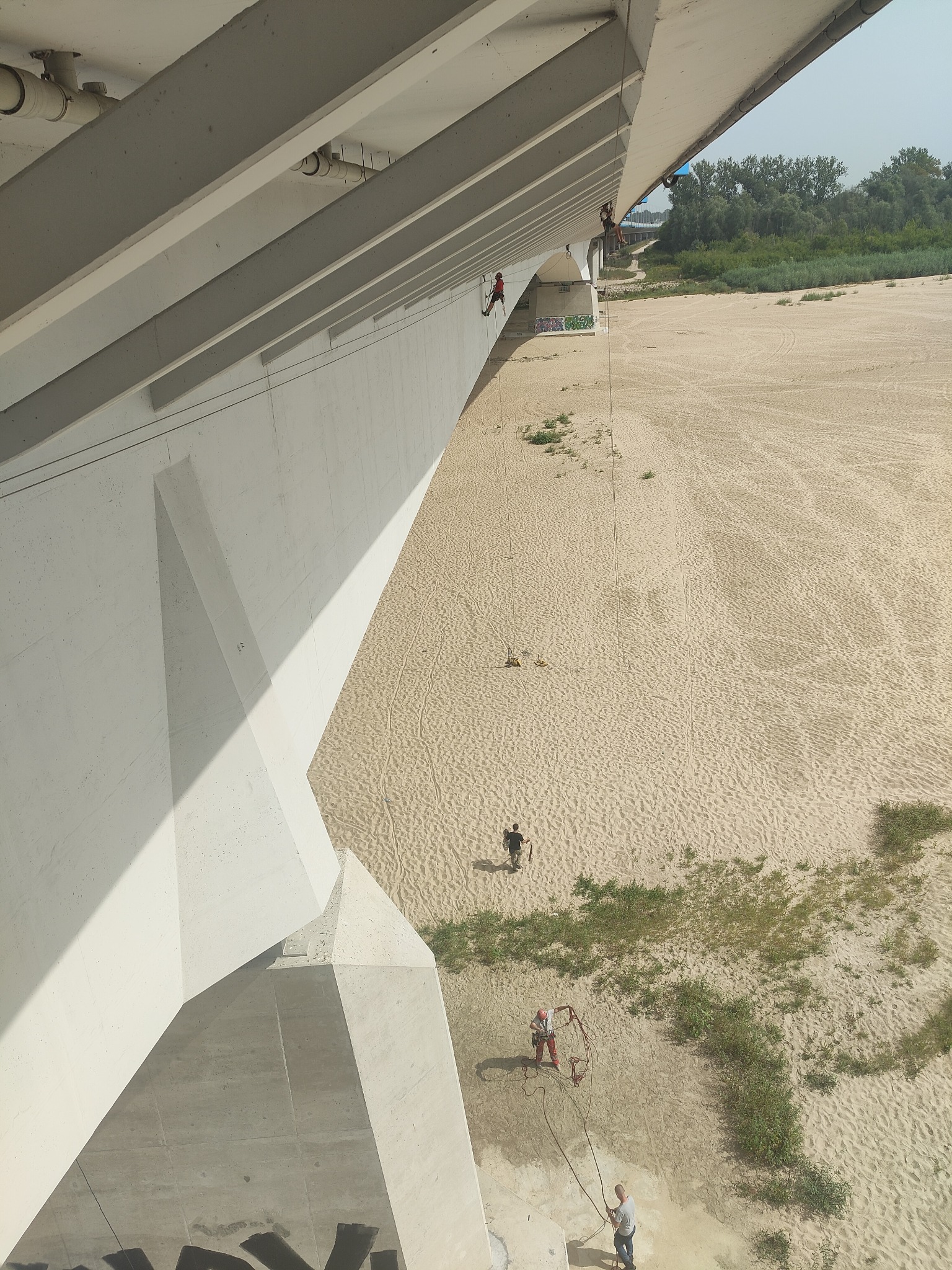 Pracownicy na linach, podwieszeni pod betonowym mostem, wykonują prace nad piaszczystym brzegiem rzeki, widoczne liny, uprzęże i elementy konstrukcji mostu.