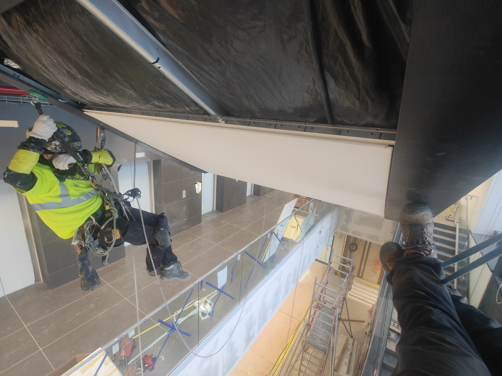 Pracownik w uprzęży linowej, zawieszony wewnątrz budynku, wykonuje prace montażowe pod sufitem, widok z perspektywy osoby stojącej na wyższej kondygnacji.