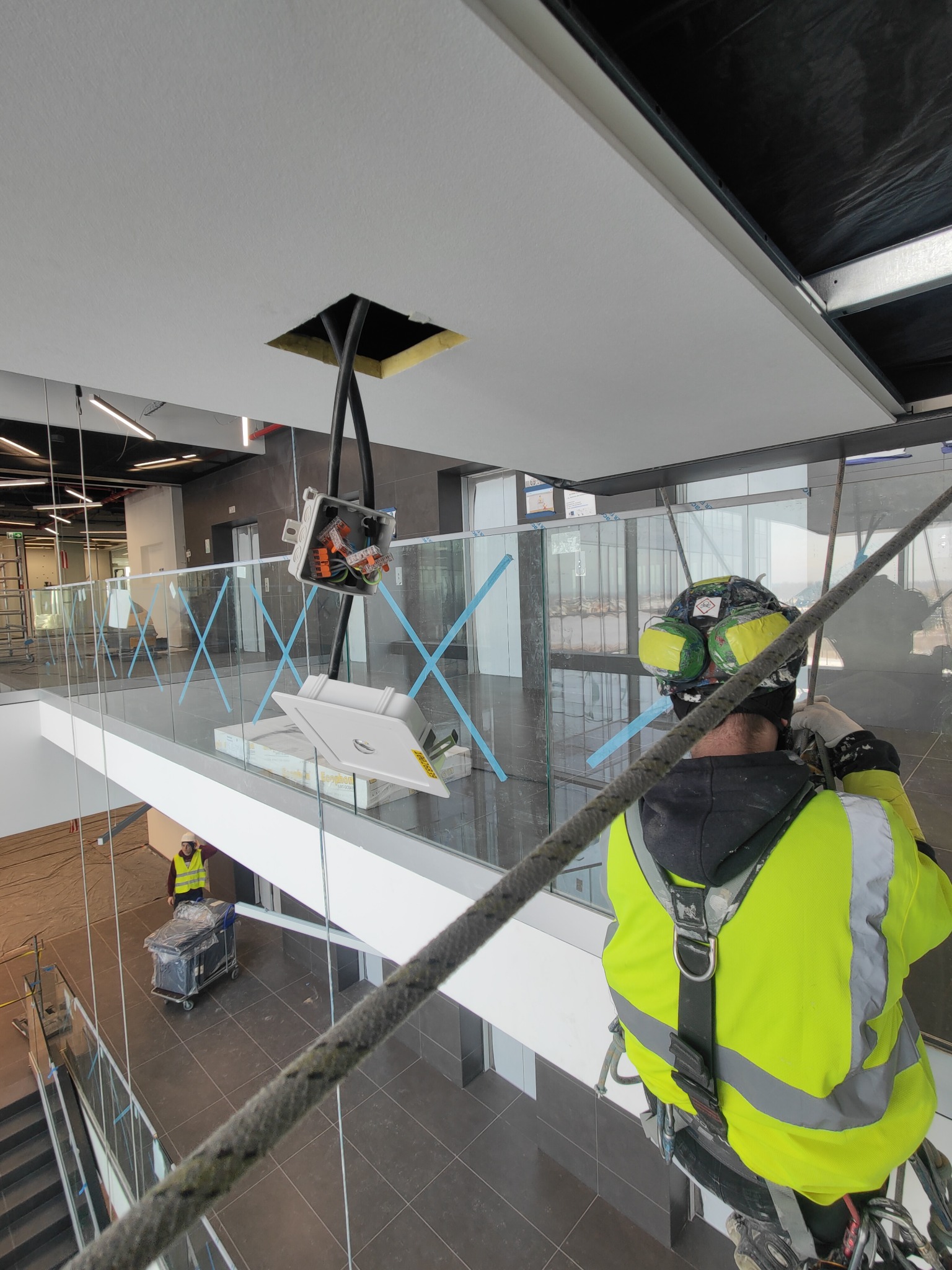 Pracownik w uprzęży wysokościowej montuje instalację elektryczną w suficie podwieszanym w nowoczesnym biurowcu z przeszkloną balustradą, widoczne przewody i puszka elektryczna, drugi pracownik...