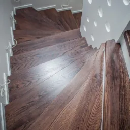 Spiralne schody z ciemnego drewna z białą, ażurową balustradą, widok z góry. Stopnie schodów z wyraźnym usłojeniem, oświetlone z góry, tworzą geometryczny wzór.