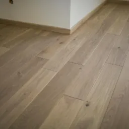 Jasne, ułożone parkietowe deski podłogowe w nowym wnętrzu, widoczne z niskiego kąta. Listwy przypodłogowe w kolorze drewna, białe ściany. Widoczny fragment grzejnika.