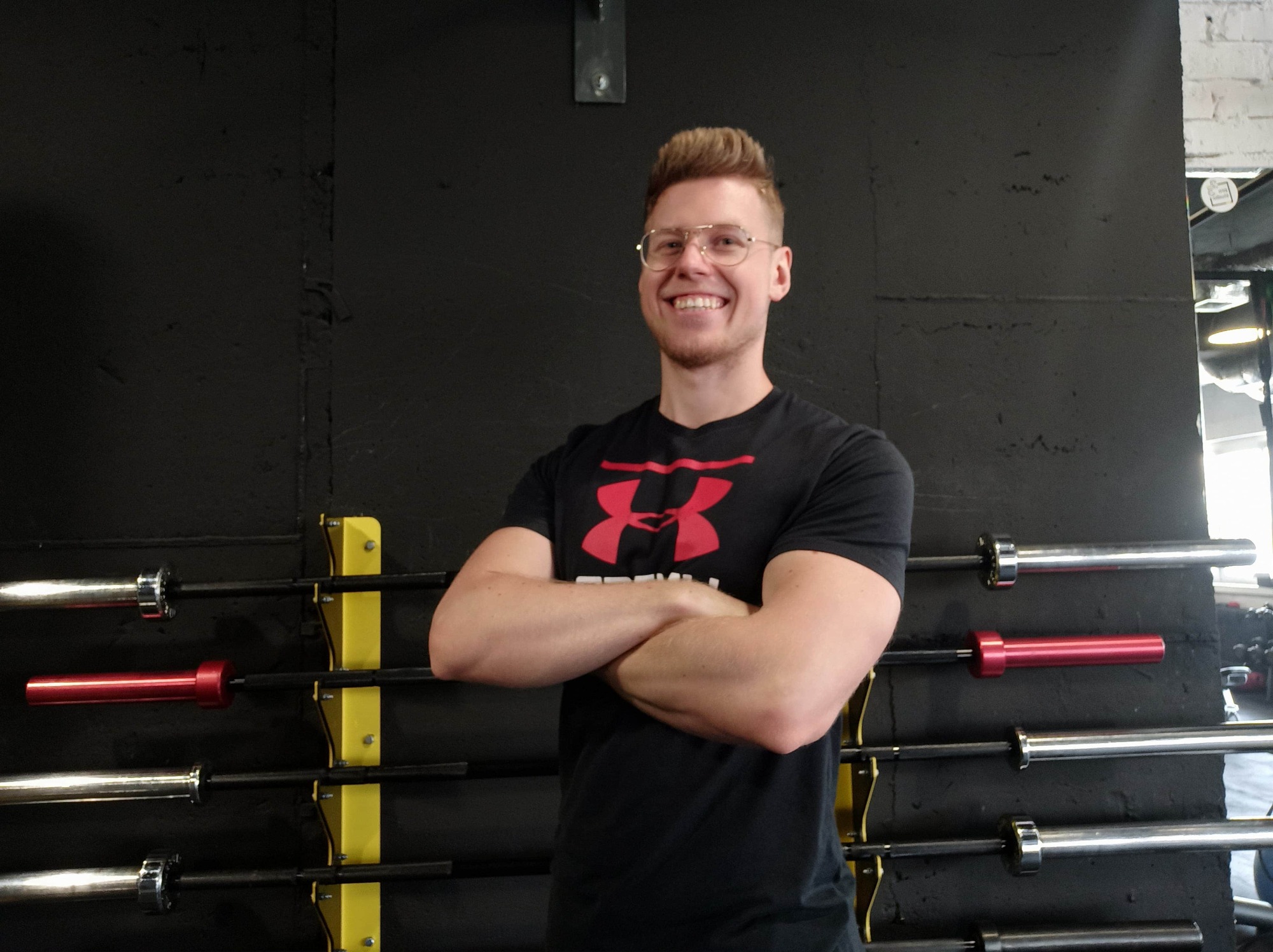 Uśmiechnięty mężczyzna w okularach, z założonymi rękami, stoi na tle ściany z uchwytami na sztangi w siłowni.