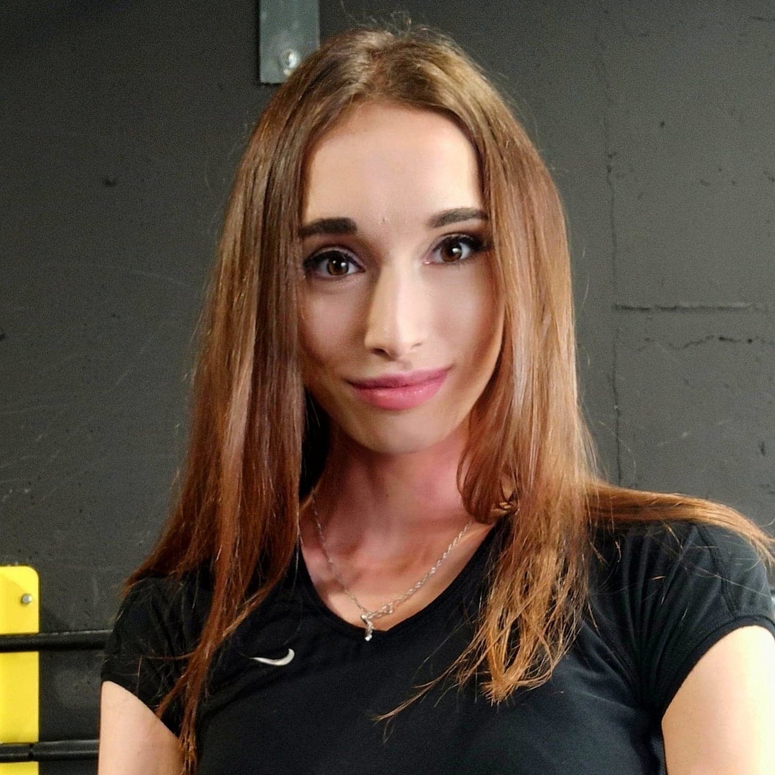 Portret uśmiechniętej kobiety z długimi, brązowymi włosami w czarnej koszulce z logo Nike, na tle szarej ściany w siłowni.